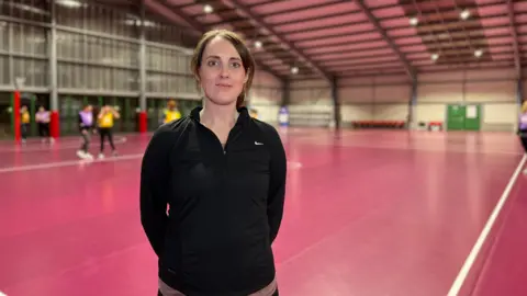 Sammy looks at the camera and her hands are behind her back. She's wearing a s black sports jacket and she has brown hair which is tied into a ponytail. Some people play netball on an indoor court behind her.