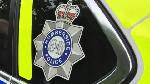 BBC The Humberside Police logo with Humberside Police in a blue circle, ER in the middle and a star and crown around it. Located on the side of a police car