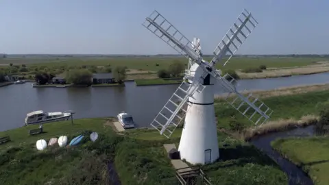 Martin Barber/ BBC Thurne Windmill complete with sails on the banks of the River Thurne, Norfolk Broads. Grassy banks, a few boats in the water, some upturned canoes on the grass banks. The land is very flat and the waterway is still. 