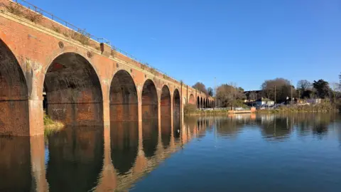 Yen Milne A sunny day with bright blue sky with a viaduct running down the left-hand side of the image. A body of water runs alongside the viaduct which is reflected on the surface
