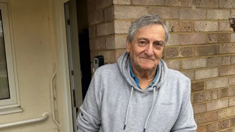 BEN SCHOFIELD/BBC Martin Swales standing outside of his home. He is wearing a light grey, hooded  sweatshirt, with two drawstrings hanging down from the hood. His hands are pushed into his sweatshirt's pockets. Martin has salt-and-pepper black and white hair, which he wears in a parting on one side. Behind him on the right are the bricks of the front of his home, as well as the very edge of a hanging basket. On the left of frame is his front door, which is open, and a window. 