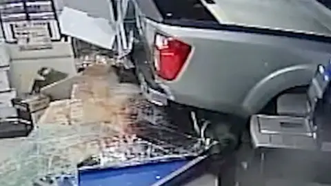 The rear of a car sits on top of the broken glass of a shopfront with debris strewn around