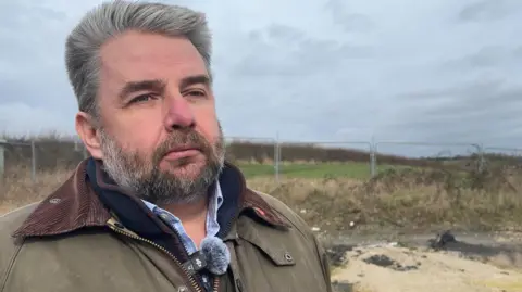 A man with slicked back grey hair and a matching beard stands in front of a fenced-off piece of green land under a grey sky. He is wearing a brown jacket over a blue shirt.