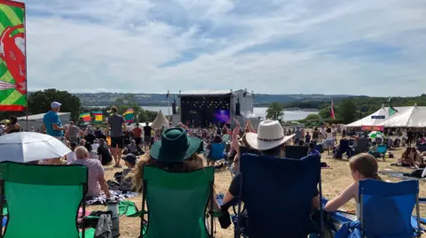 Three people sat in deckchairs, facing a stage. You can see the tops of their heads, with two wearing hats. Crowds of people are in front of them, cheering on performers on a large stage in the distance.