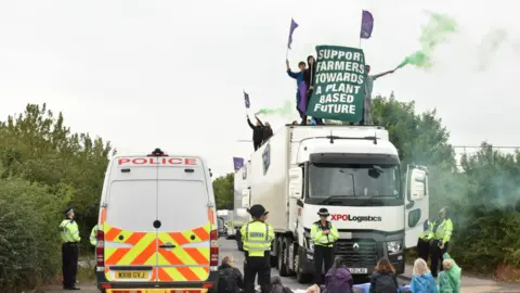 A police van and a lorry stand surrounded by protesters, some of them sitting on the ground, as well as police officers. People standing on top of the lorry are holding coloured flares and a banner reading "Support farmers towards a plant based future."