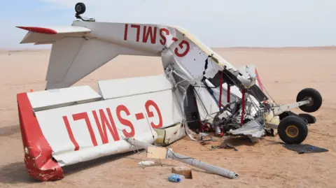 Leandrea Mouers/Erongo A small white plane with red detailing, which has crashed in a sandy desert. The plane is upside down and has suffered significant damage, mostly to its cockpit.