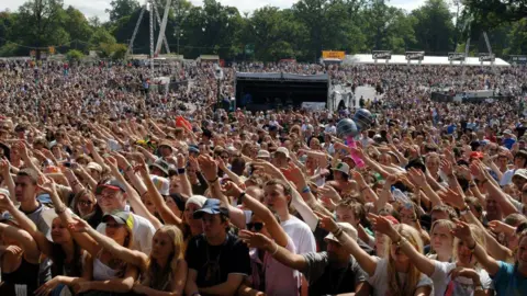 PA Media A crowd of hundreds of people wave their arms in unison. The weather is hot and many people are wearing hat and sunglasses. The crowd is extensive and a beer tent is in the far distance.