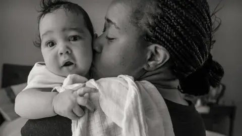 Naturally Tiwa Skincare Shalom and Joshua Lloyd, a woman holding a baby, the picture is in black and white and the woman is kissing the boy, he is looking at the camera and the woman is looking at the baby