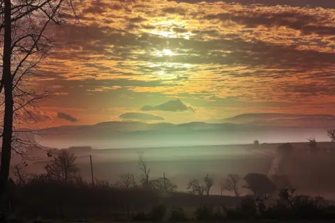 Leonie66/BBC Weather Watchers The sun is shining through clouds in an orange sky above mist shrouded fields at Kelso.