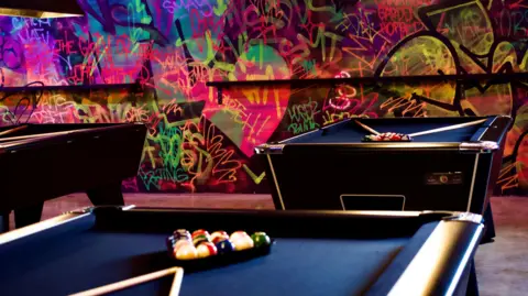 Three pool tables in a room, with graffiti art on the walls, the pool tables are blue with ball and cues on them. 