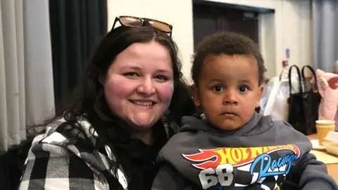 JOHN FAIRHALL/BBC A woman in her mid-20s with shoulder-length dark hair and sunglasses on top of her head is smiling at the camera. She is wearing a white and black checked top. On her lap is her toddler son who is wearing a grey Hot Wheels Racing hoodie.