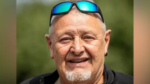 Steve Leadbetter, smiling and looking at the camera, with a grey goatee and short grey hair, with sunglasses on his head, he has an earring in his left ear, and is wearing a black top.