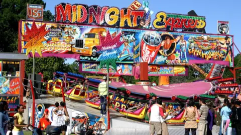 A colourful funfair ride with people standing around in front of the ride. 