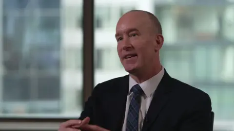 A man in a dark suit looks to the left of the camera, with his hands clasping in motion. He is wearing a white shirt and blue and black chequered tie. The man has short grey hair and is sitting in an office, with the windows in the background out of focus. 