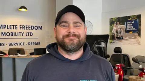 Ben Molloy, the manager at Complete Freedom Mobility in Sutton-in-Ashfield, smiling into the camera with mobility scooters in the background.