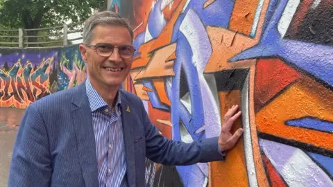 A smiling man wearing a blue suit and glasses puts his hand on a newly decorated wall.
