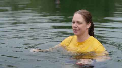 A woman with brown hair in a ponytail is wearing a yellow T-shirt and floating in a lake. The water looks green.