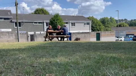 Three men sit on a park bench on Wilmot Gardens in Plymouth. Several terrace houses with grey walls are behind them along with a few trees.