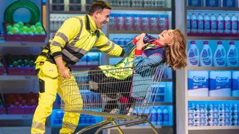 Pamela Raith A woman in a supermarket uniform of stripy blue shirt flings her blonde hair back while sitting in a supermarket trolley being pushed by a man in hi-vis workwear. Both are smiling. She is holding a can of Fosters lager.