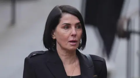 Sadie Frost, a woman with short, straight black hair, looks ahead with a blank expression. She is wearing a black jacket above a black top, with a gold necklace.