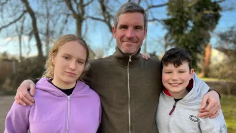 Stuart Woodward/BBC Neil Duckett, wearing a dark green zip-up fleece, stood between his daughter Toula, wearing a purple hoodie, and Joe who is wearing a light grey hoodie. Neil has his arms around his children's shoulders.