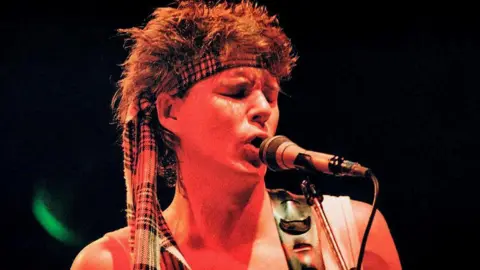 Getty Images Stuart Adamson of Big Country singing into a microphone in a photo from the eighties. He has a spikey mullet and a tartan scarf tied round his head
