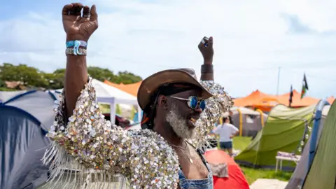 A man wearing a wide-brimmed hat, a sequin-crusted jacket, dungarees and sunglasses holds his arms aloft in a celebratory pose, among a field of tents.