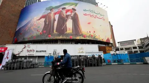 Reuters A man on a motorcycle looks at a large billboard featuring Iran's new Supreme Leader Mojtaba Khamenei, and late Supreme Leaders Ayatollah Ali Khamenei and Ayatollah Ruhollah Khomeini, amid the U.S.-Israeli conflict with Iran, in Tehran, Iran, March 12, 2026.