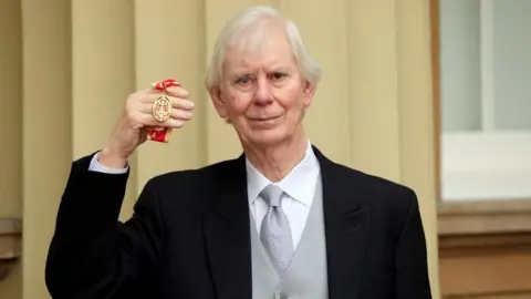 PA Media Sir Andreas Whittam Smith wears a black suit with a grey waistcoat and tie. He has white hair and is standing in front of what looks like a pillar and holding up a gold medal on a red ribbon. 