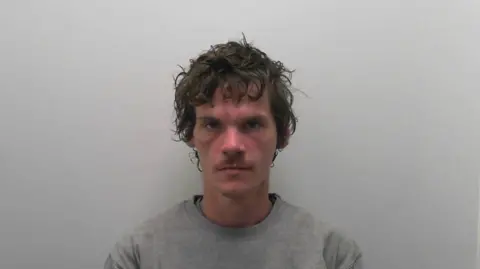 Daniel Bell in a police mugshot. He has medium length brown hair and a small brown moustache and is wearing a grey sweatshirt