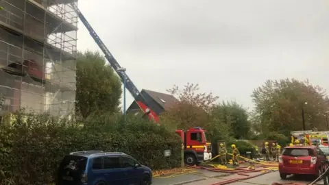 Fire engine outside tower block, with firefighters and hoses nearby. There is also a red and blue car parked nearby.