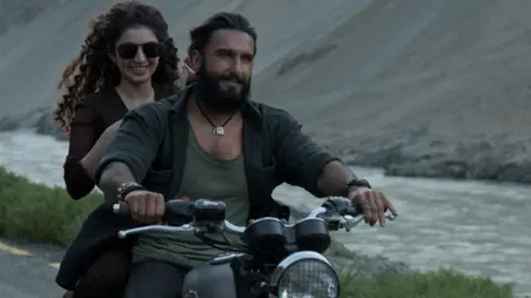 A scene from Dhurandhar movie in which actor Ranveer Singh, wearing an unbuttoned shirt over a vest, and sporting a beard, is riding a motorbike, next to a river. Sitting on the pillion is actress Sara Arjun, wearing a black kurta and sporting aviator sunglasses