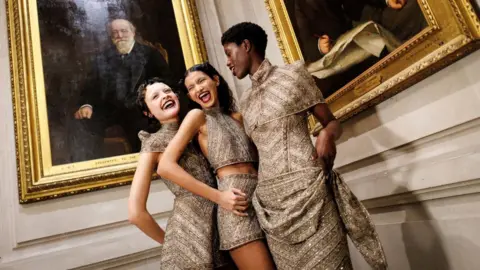 Tolga Akmen/Shutterstock Three models in sparkly gold dresses stand in front of two portraits of men