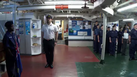 A naval officer and group of cadets stand to attention on board a warship