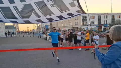 Laurie Argent with his arms in the air as he nears a red ribbon finish line in Worthing. There is a small crowd of people on the pavement near him and a black and white finish flag being held up in the foreground.