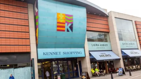 An external picture of the Kennet Shopping centre with a large blue sign, a cafe nero and laura ashley.