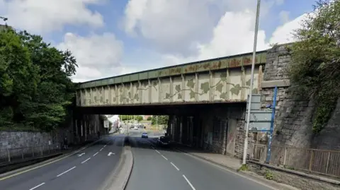 A white and green metal railway bridge crossing over four lanes of traffic near the Pennycomequick roundabout in Plymouth. There are trees on either side of the road and two cars crossing under the bridge. It is a cloudy but sunny day.