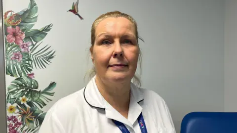 Ellen Knight/BBC Van Duivenbooden pictured looking into the camera and smiling slightly. She is wearing a white collared shirt with a dark blue trim on the collar. She has blonde hair, which is tied back into a ponytail, and is also wearing a dark blue lanyard. The room she is stood in has blue-grey walls, and has a mural painted on the wall of tropical flowers and leaves, with a hummingbird. 