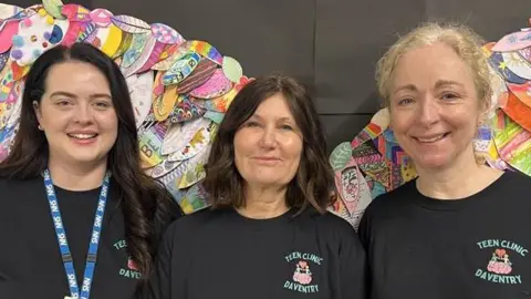 Three women stood side-by-side in black T-shirts. All are looking at the camera and smiling. Behind them is a colourful display.