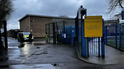 Modern brown brick school building with a police officer standing outside and a police van
