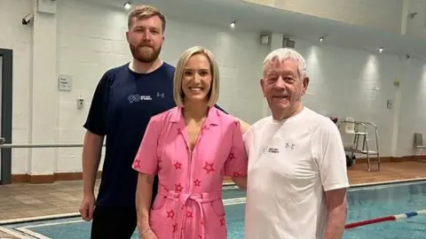 BBC Kay Crewdson is standing at the side of an indoor swimming pool next to Ken and his grandson Sam. Both of the men are wearing T-shirts and their swimwear