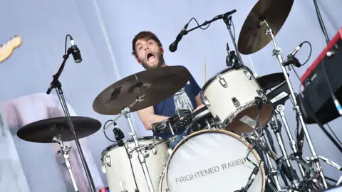 A man sits behind a drum set on a stage. He had red ear plugs in his ears. shaggy beard and hair. he also has his mouth open as if he was singing along with a rock song.