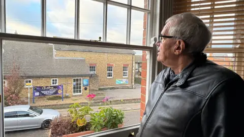 Mike Bromberg with short white hair and glasses, wearing a grey zip-up jacket, looking out of a sliding window at a brick-built church on the other side of the road. A grey car is passing the church.