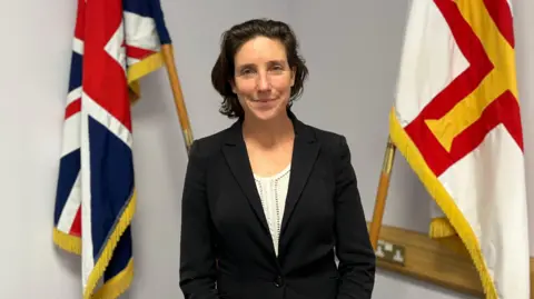 Lindsay de Sausmarez - A woman with short dark hair smiling at the camera. She is wearing a white blouse with a black blazer over it. There is a Guernsey flag and a Union flag behind her.