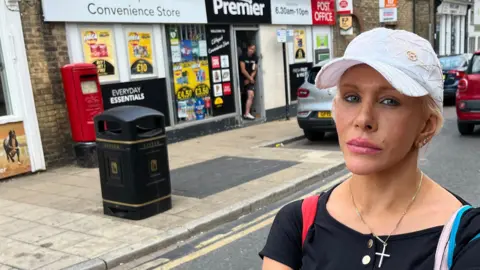 John Devine/BBC Juliet Carter is standing in a street in a town looking at the camera. She has short, blonde hair and is wearing a white baseball cap. She has a black T-shirt on and a gold necklace. Shops, cars, a litter bin and a post box can also be seen in the photo.