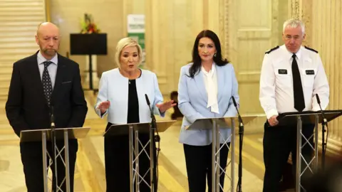 PA Media Four people stand behind lecterns with microphones, addressing a press briefing in a large, ornate indoor hall with marble walls and tall columns. All are dressed in formal or professional attire, and one person on the right wears a uniform with a visible police badge.
