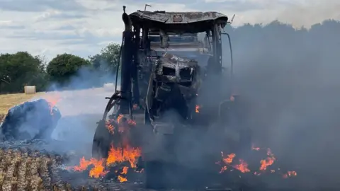 Judith Taylor A blackened and burning tractor in a field. Its structure has warped in the fire, which is still blazing around its tyres with smoke rising. Behind is yellow stubble, two round straw bales can be seen, trees and blue sky.