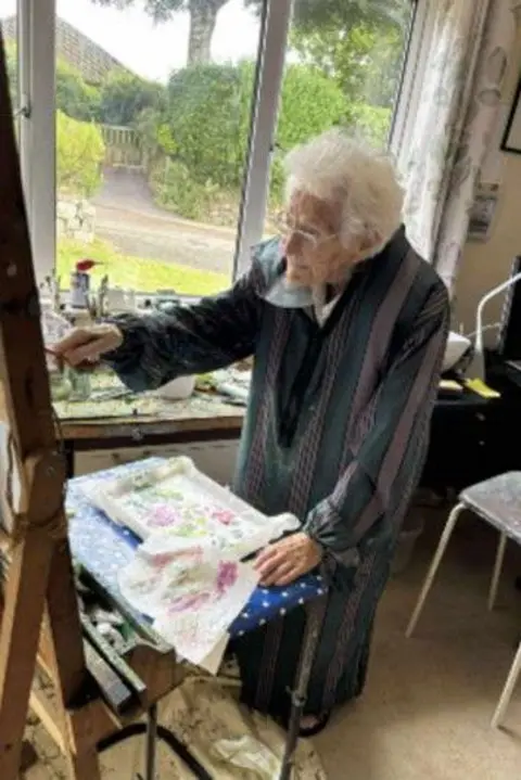 Penny Lees 107-year-old Mollie Silver in her house painting some artwork. She is wearing a green and brown overall and is putting a brush on the canvas. She has a tray with a palette of paints on it propped up on a stool in front of her.