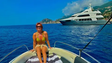 Facebook Kershaw is sat down on the front of a boat facing away from the sea. Behind her is a large yacht and a coastline.