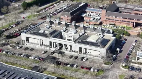 An aerial view of Milton Keynes Magistrates' Court with a prominent light-coloured building in the foreground. The building has a rectangular footprint, several roof-mounted solar panel arrays, and multiple skylight-style roof structures. Parking areas with numerous cars surround the complex, and additional brick buildings are connected behind it. Roads, trees, landscaped areas and more parked vehicles are visible around the site.